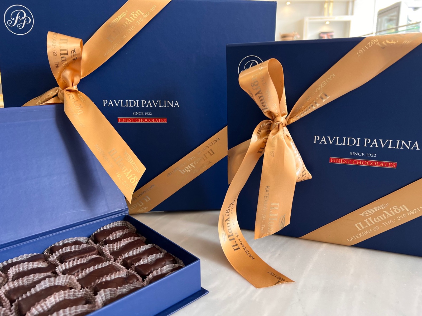 Ανοιχτό μπλε κουτί δώρου Pavlidi Pavlina με μάντολες, δίπλα σε κουτιά με χρυσή κορδέλα